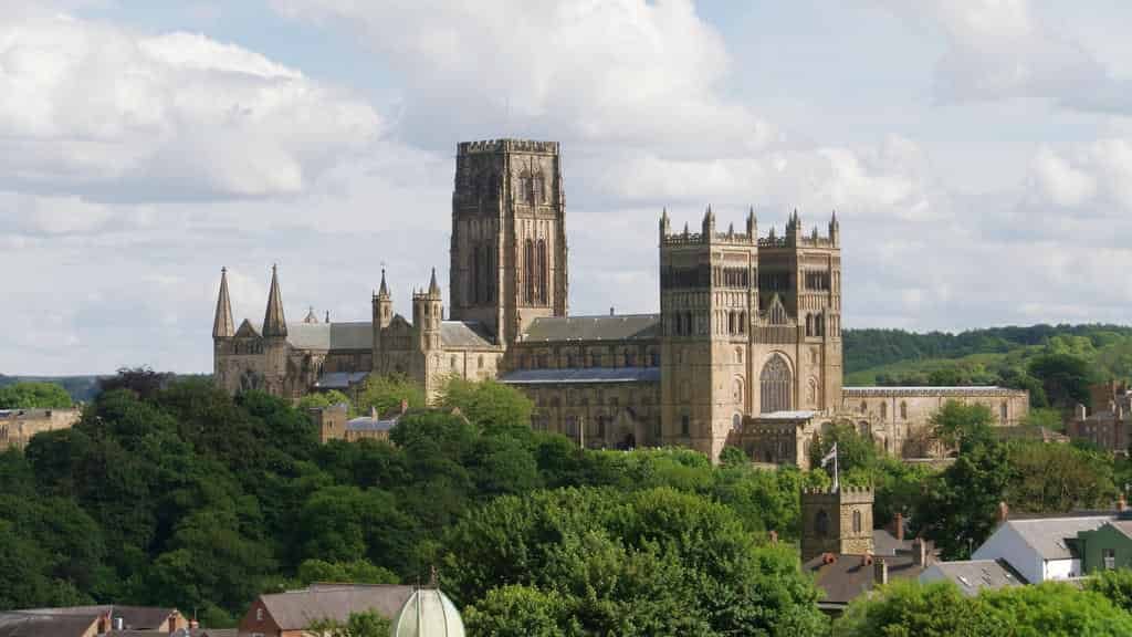 Distant panoramic view of the large, historic, stone Durham Cathedral with its central tower, rising above a dense canopy of green trees on a hill.