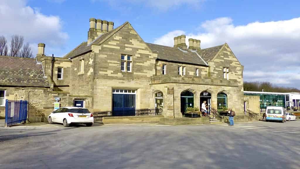 The historic stone facade of Durham Railway Station with traditional architecture, chimneys, and a small forecourt on a sunny day.