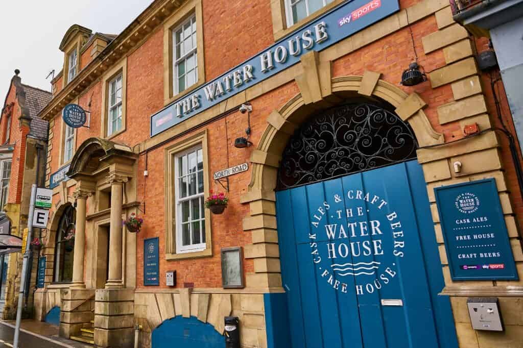 Exterior of The Water House pub in Durham, a red brick building with an arched stone entrance and a large, blue wooden door stenciled with white text advertising cask ales and craft beers.