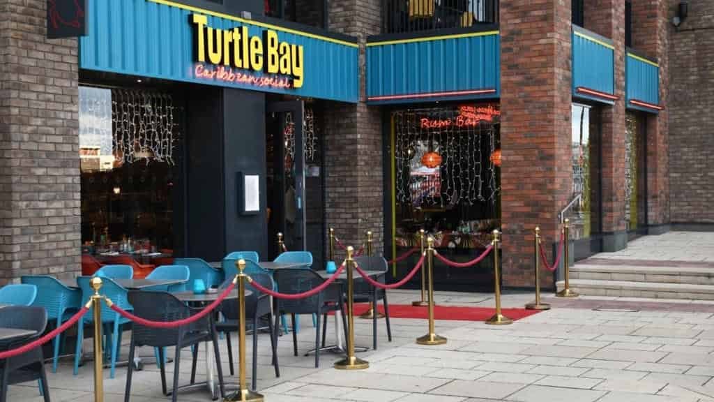 Exterior of the Turtle Bay Caribbean Social restaurant, a red brick building with a bright turquoise sign. The foreground shows an outdoor seating area separated by red velvet ropes and gold stanchions.