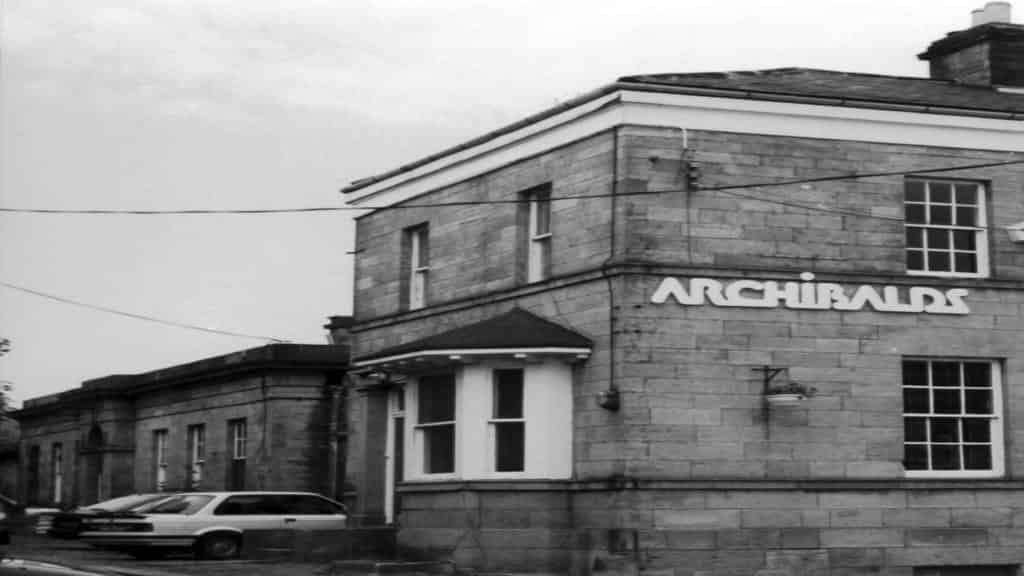 History of Archibald's at Gilesgate Station: A Black and white photograph of the stone building "Archibalds" in Gilesgate, Durham. The name ARCHIBALDS is clearly visible in white letters on the facade. A corner bay window is on the ground floor, and parked cars are in the foreground.