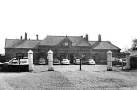 Black and white photograph of the old Durham Elvet train station building. The Victorian-style building has a central clock. In the foreground, a large forecourt is visible with several mid-20th-century cars and two prominent stone entrance pillars.