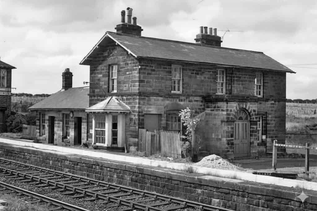 Shincliffe's Lost Railway Stations: A Black and white photo of a two-story stone railway station house with a bay window, standing on a platform beside the tracks.