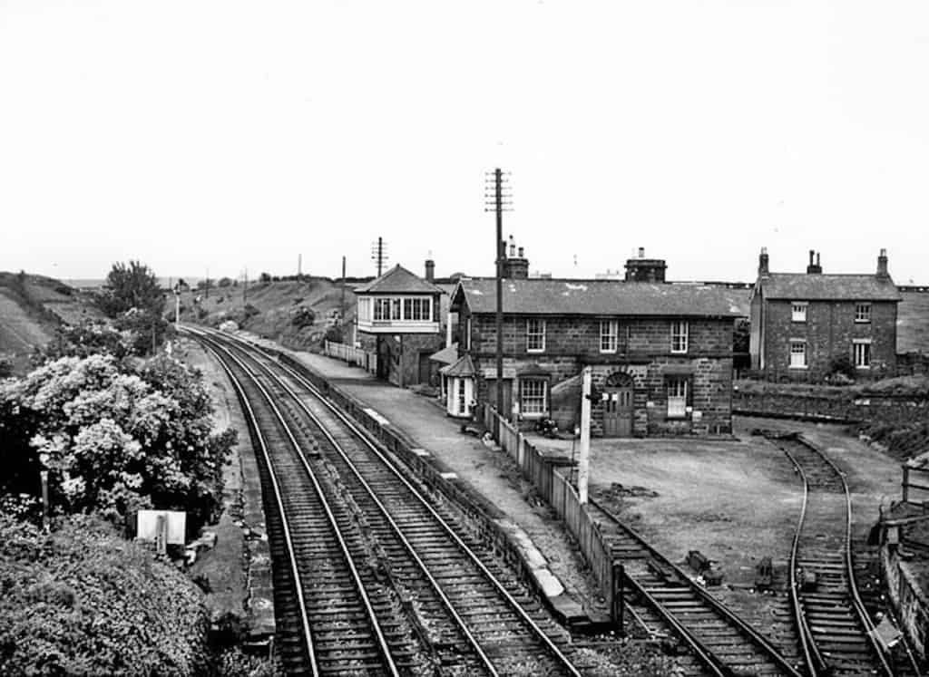 Historical black and white image of a railway station. Two sets of tracks curve to the left, passing a two-story stone station house and a wooden signal box. A small siding is visible to the right.